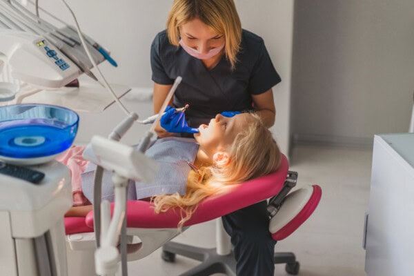 A bright, cheerful dental office setting features a young girl comfortably seated in a pink dental chair while a caring dental professional performs an examination.
